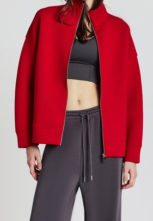 Femme portant une veste rouge zippée sur un soutien-gorge de sport noir et un pantalon gris foncé à cordon, debout devant un fond uni.