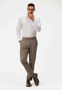 Witte blouse met lange mouwen en zwarte knopen, gecombineerd met lichtbruine broek en bruine schoenen. Model staat met één hand aan de kin.