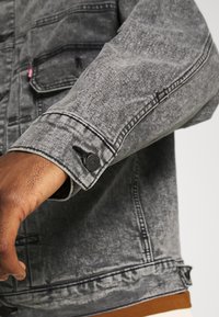 Gros plan sur une main à la peau foncée portant une veste en jean noire délavée avec un poignet boutonné et des coutures visibles de la poche.