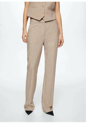 Pantalon beige sur mesure taille haute porté avec un gilet assorti et des chaussures noires à bout pointu sur une personne avec un fond blanc uni.