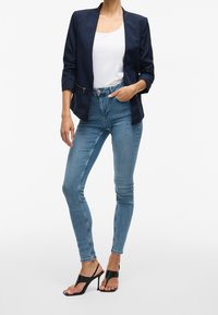 Sotilaallinen bleiseri, jossa on slim fit -leikkaus, varustettuna vetoketjullisella taskulla, yhdistettynä korkeavyötäröisiin sinisiin skinny-farkkuihin ja mustiin korkokenkiin.