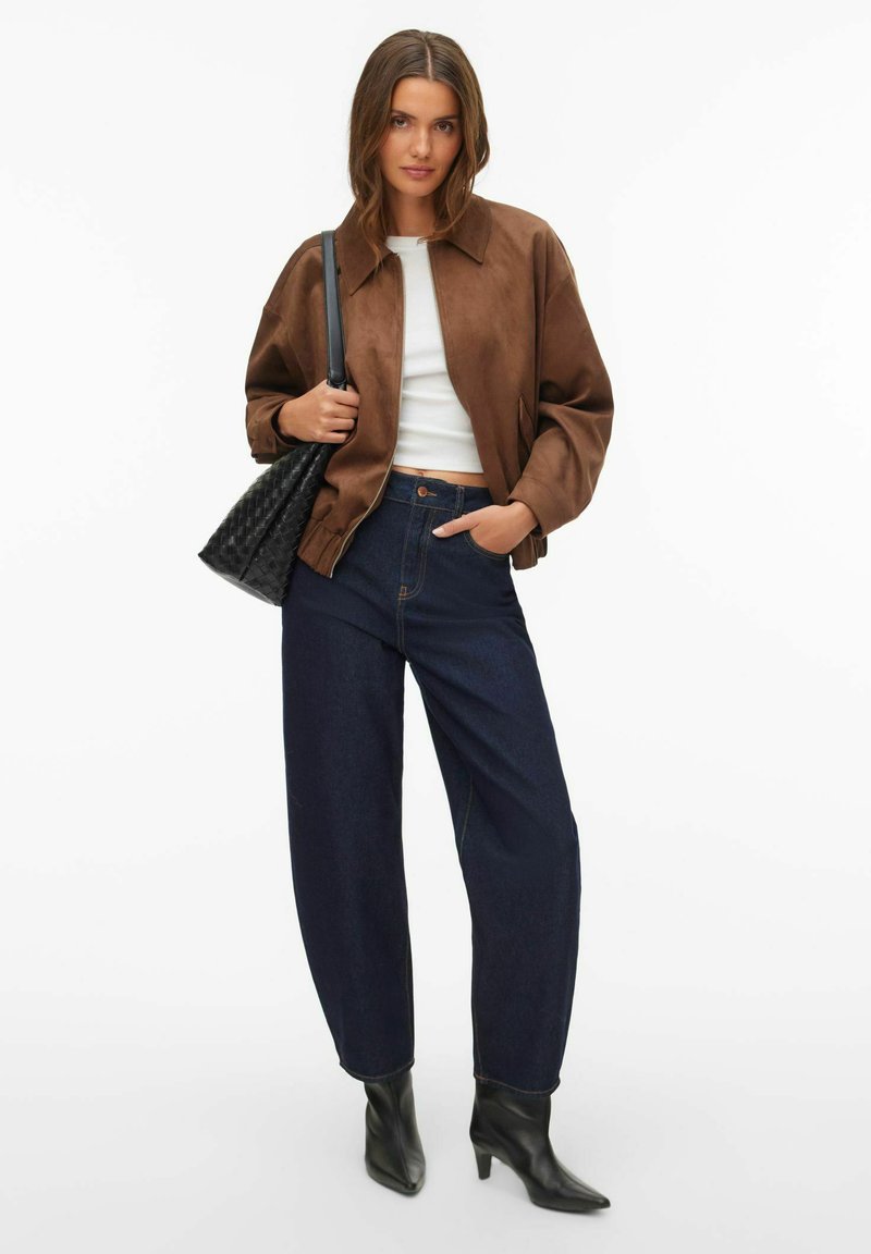 Smeđa bomber jakna od brušene kože, bijela majica, tamnoplave traperice širokih nogavica visokog struka, crne čizme s oštrim vrhom do gležnja i teksturirana crna torba.