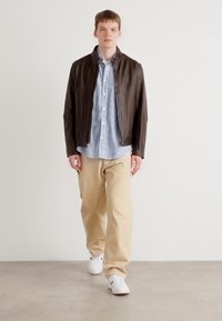 Brun lærjakke over en blå og hvit stripete skjorte, sammen med beige bukser og hvite sneakers, mot en nøytral bakgrunn.