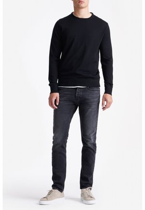 Man in een zwarte ronde hals trui, vervaagde zwarte jeans en beige sneakers die tegen een witte achtergrond staat.