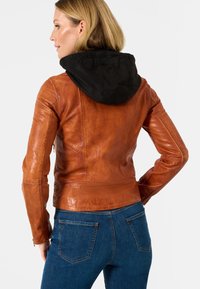 Brun læderjakke med sort hætte, tætsiddende design, synlige syninger og lynlås, parret med blå denim jeans.