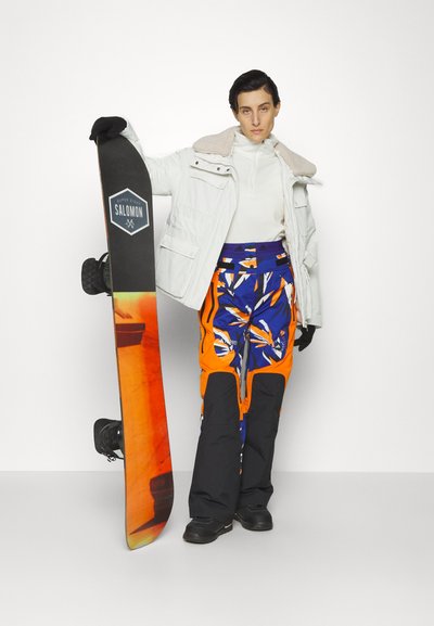 Snowboard met een oranje en blauwe gradientontwerp, vastgehouden door een persoon in een witte jas en geblokte sneeuwbroek. Zwarte handschoenen en boots zichtbaar.