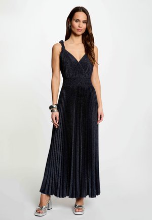 Kvinde iført en ærmeløs, plisseret, mørkeblå maxi-kjole med snoede skulderstropper og sølvfarvede platformhøje hæle på en hvid baggrund.