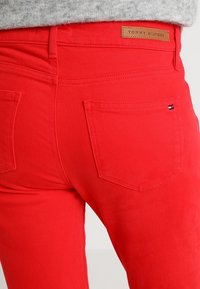 Jeans de mezclilla roja con un diseño ajustado, bolsillos traseros y una etiqueta de marca de cuero en la cintura. La textura parece suave y duradera.
