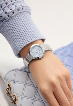 Montre argentée avec un cadran rond, un cadran bleu clair et un bracelet en argent texturé. Portée sur une main avec une manche tricotée bleu clair et un sac matelassé.