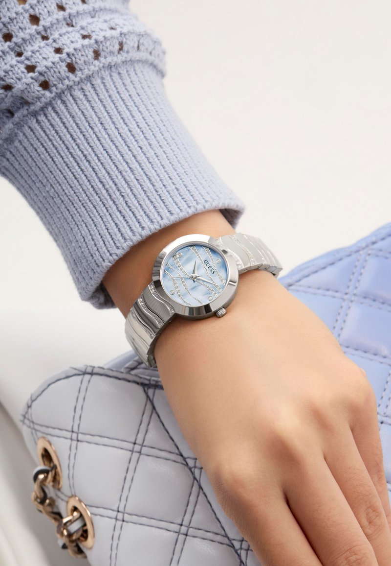 Orologio silver con quadrante rotondo, dial azzurro chiaro e cinturino in argento testurizzato. Indossato su una mano con una manica in maglia azzurra chiara e una borsa trapuntata.