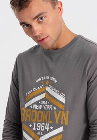 Graues Langarmshirt mit einem bedruckten Brooklyn-Motiv in Weiß und Gold, rundem Ausschnitt und weicher Textur.