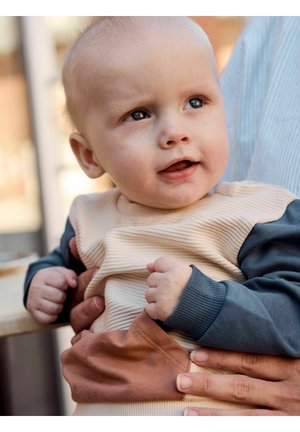 Baby med blå øjne og lyst hår iført en trøje med striber og blå ærmer sidder i en voksens arme og kigger let opad med et smil.