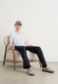Camicia azzurra chiara a maniche corte, pantaloni neri, cappello grigio e sneaker beige. Seduto su una sedia curva di legno con un cuscino crema.