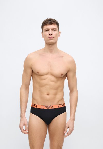 Μυώδης άνδρας στέκεται μπροστά σε λευκό φόντο φορώντας μαύρα εσώρουχα με πορτοκαλί ζώνη μέσης που φέρει το λογότυπο EA και έμβλημα αετού.