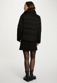 Sorth quilted pufferjakke med høj krave, prydet med et subtilt præg mønster, parret med en sort plisseret nederdel og kraftige sorte støvler.