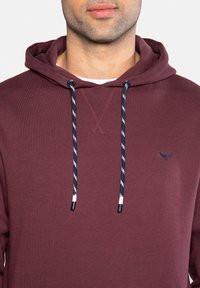 Homme portant un sweat à capuche bordeaux avec des cordons de serrage noirs et blancs et un petit logo brodé sur la poitrine.
