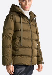 Doudoune verte olive avec capuche, présentant un zip sur le devant et deux poches zippées sur les côtés. Fabriquée en tissu matelassé et lisse avec des sangles ajustables.
