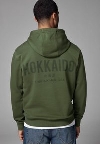 Sweat à capuche vert olive avec un grand imprimé noir "HOKKAIDO" au dos, accompagné d'un texte plus petit en dessous. Texture douce, capuche ajustable.