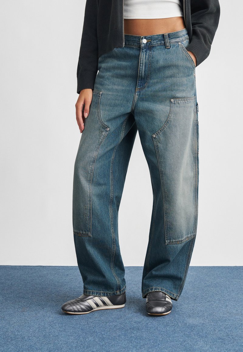 Lyseblå denimjeans med løs pasform, der har store forlommer, synlige syninger og en falmet vask. Kombineret med sorte atletiske sko.