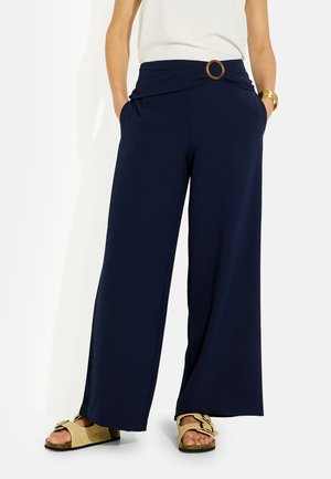 Femme portant un pantalon large bleu marine avec une boucle de ceinture circulaire, un haut blanc, un bracelet doré et des sandales beiges avec des boucles.