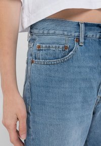 Närbild av en person som bär ljusblå denimjeans med en vit kortärmad skjorta, där framlommen och en del av handen syns.