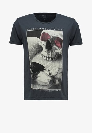 T-shirt grise avec un graphic de crâne, manches courtes et encolure ronde. Porté avec des shorts en denim retroussés et des baskets blanches, tenant une casquette noire.