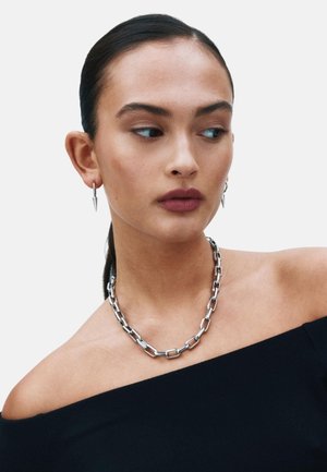 Ogrlica iz srebrne verižice s pravokotnimi členki in uhani v obliki bodic, ki jih nosi model z temnimi lasmi in nevtralno ličilo na enostavni ozadju.