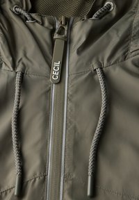 Giubbotto a vento verde oliva con zip frontale, cappuccio con cordino e fodera testurizzata. Presenta un tirazip con logo e cuciture in contrasto.