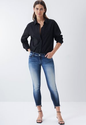 Chemise noire à boutons, jeans skinny dégradé bleu clair à bleu foncé avec ourlet brut, et sandales noires à lanières. Couture détaillée sur le pantalon.