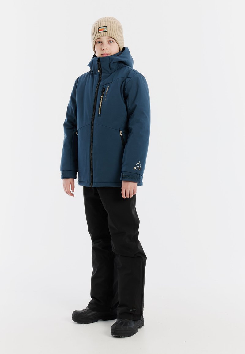 Marineblaue Softshell-Jacke mit hohem Kragen, Reißverschlusstaschen und einem Berg-Logo; kombiniert mit schwarzen Hosen und robusten schwarzen Stiefeln.