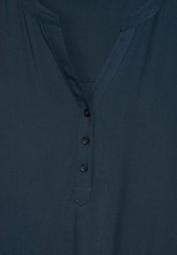 Camicetta blu scuro con scollatura a V e tre bottoni neri allineati verticalmente sulla patta frontale.