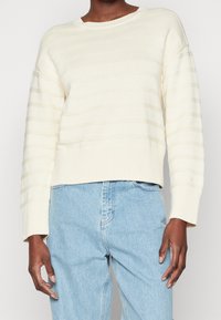 Suéter de punto color crema con rayas horizontales, cuello redondo y dobladillo acanalado. Lleva puesto un pantalón de mezclilla azul claro.