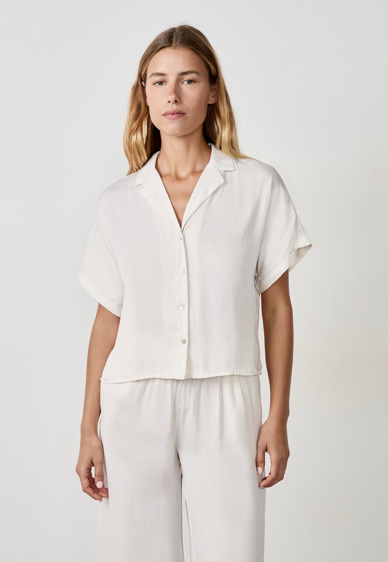 Camicia a maniche corte, con bottoni, di colore crema chiaro, realizzata in un tessuto morbido. Presenta un colletto a punta e una vestibilità rilassata con lunghezza cropped.