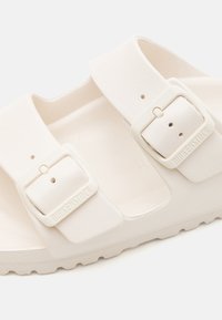 Hvite Birkenstock sandaler laget av mykt materiale, med to justerbare remmer med spenner, en teksturert såle og en riller på sålen.