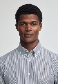 Randig skjorta med knappar i blått och vitt, med krage, vita knappar och en liten röd logotyp på vänster bröst. Slät tygkvalitet.