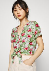 Top de flores curto em cinza claro com flores de hibisco rosa e folhas verdes, apresentando um nó amarrado na frente e mangas curtas.