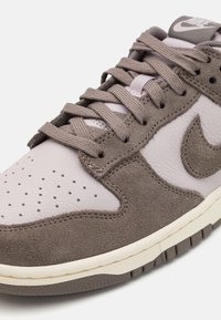 Hnedé a svetlošedé tenisky vyrobené z kože a zamatu, s perforovaným špiceom, šnurovaním a bielou medzi podrážkou. Obsahuje logo Nike swoosh.