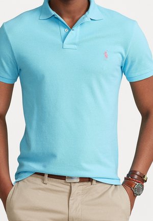 Polo bleu clair en tissu texturé, avec une patte de boutonnage à trois boutons et un petit logo rose brodé sur le côté gauche de la poitrine.