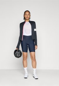 Sort cykeljakke med lange ærmer og lyserøde detaljer, hvid mesh undertrøje, marineblå cykelshorts, hvide sokker, holder en sort hjelm.
