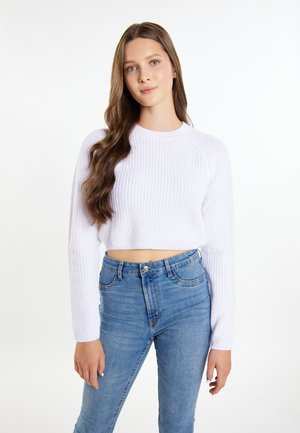 Ung kvinde med langt bølget brunt hår iført en hvid, kort strikket sweater og højtaljede blå jeans, stående mod en ensfarvet hvid baggrund.