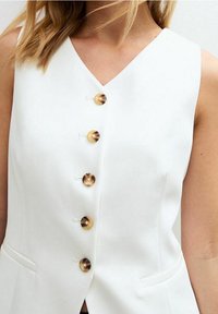 Nainen, jolla on päällään hihaton valkoinen toppi, jossa on V-pääntie ja viisi suurta ruskeaa kilpikonnakuosisia nappia keskitettynä eteen.