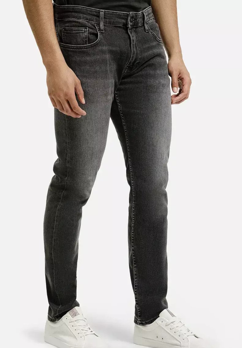 Mężczyzna w dopasowanych czarnych spodniach z lekkim przetarciem i białych sneakersach, stojący z rękami swobodnie wzdłuż ciała na jednolitym tle.