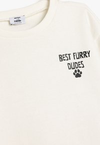 Sweatshirt de couleur crème avec un col côtelé. Présente le texte noir "MEILLEURS CHIENS POILUS" et un graphique de patte à l'avant. Texture douce.