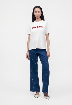 Kvinde stående iført en hvid t-shirt med teksten "Côte d'Azur", blå mønstrede jeans og hvide spidse hæle mod en ensfarvet baggrund.