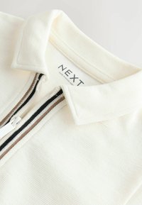 Haut en tissu côtelé blanc avec un col et une fermeture éclair avant présentant des rayures noires et marron, étiqueté "NEXT" à l'intérieur du col.