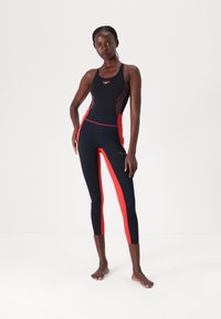 Black athletic bodysuit with red accents and geometric lines, paired with fitted black leggings featuring red side panels and a high waistband.