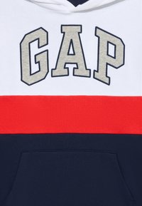Felpa con cappuccio caratterizzata da un design a spacco: parte superiore bianca, striscia orizzontale rossa e parte inferiore blu navy. Logo "GAP" prominente in beige.