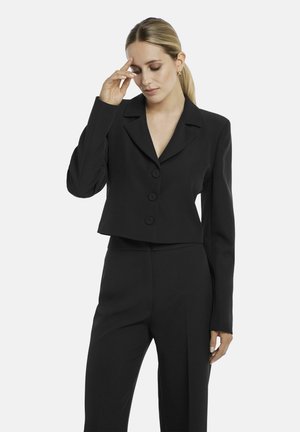 Woman with blonde hair in a low ponytail wearing a black cropped blazer and matching high-waisted pants, touching her temple with one hand.