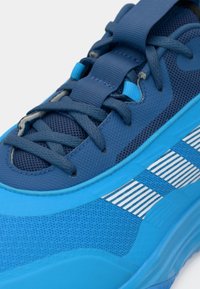 Blå athletisk sko med mesh-overside, teksturert detaljering og hvite stripete aksenter. Har flate lisser og en dempet såle.