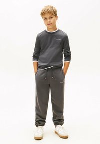 Junge steht mit Händen in den Taschen, trägt einen grauen Tommy Hilfiger Sweatshirt und passende Jogginghose mit weißen Sneakers vor weißem Hintergrund.